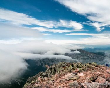 "Ascending Pamola" - A hiker in a blue jacket approaches the top of Pamola Peak from the Dudley Trail as the wind whips the clouds about.