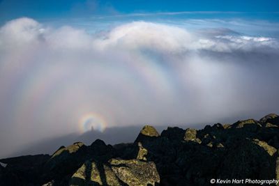 A small circular rainbow containing the shadow of the photographer is seen on clouds below the rocks of the summit, surrounded by a larger fog bow.