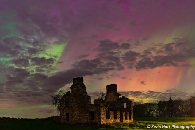 Aurora colors including green, yellow, magenta, purple, and orange fill the sky among clearing clouds above an abandoned building.