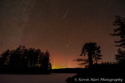 Three geminids streak through the sky while the red aurora dances below, framed by silhouetted trees. The frozen lake is tinted pink. Vermont.